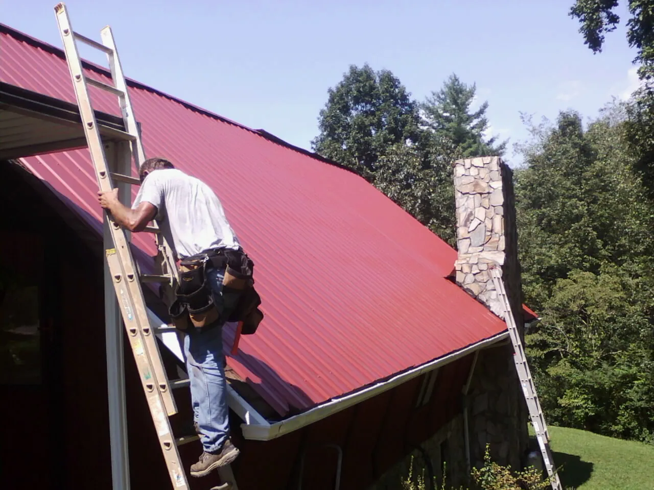 Roofer on a ladder beside a red metal roof and stone chimney — Soffit & Fascia Repair by Roofing Brothers in West Islip